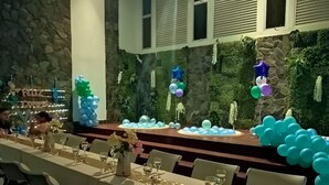 Una larga mesa de comedor dispuesta con sillas, globos y un muro de piedra de fondo.