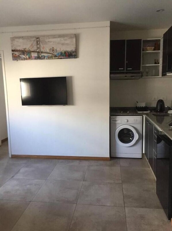 Eine kompakte Küche mit integriertem Waschmaschine, einem flachbildfernseher an der Wand und einer kleinen Arbeitsfläche.