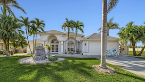 A two-story house with a white exterior, a stone feature in front, and palm trees around. - Villa Palm Garden heated Pool (Cape Coral)