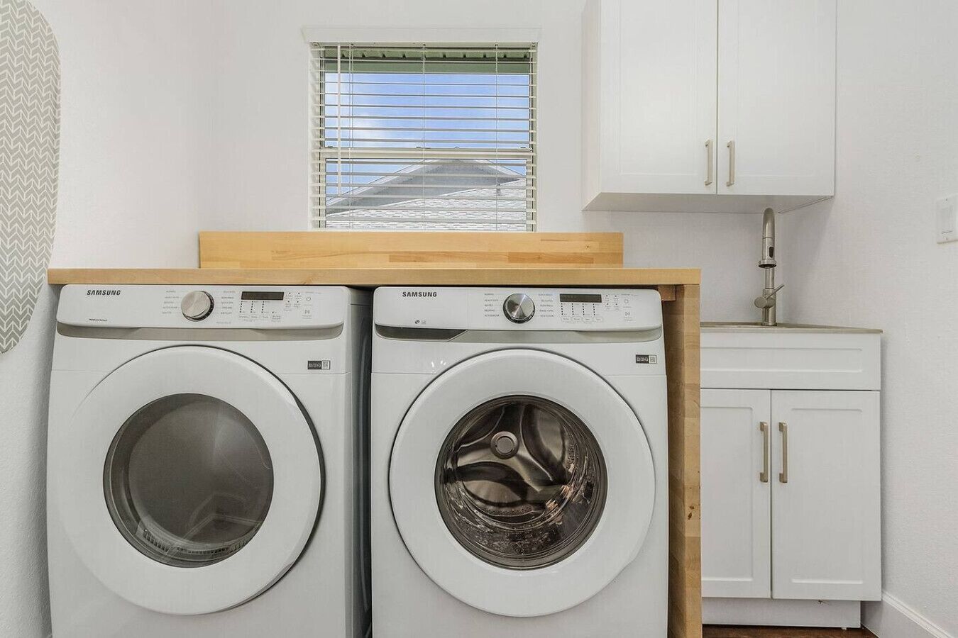 Ein Waschraum mit zwei Samsung-Waschmaschinen, einem Fenster mit Jalousien und einem weißen Schrank mit Waschbecken.