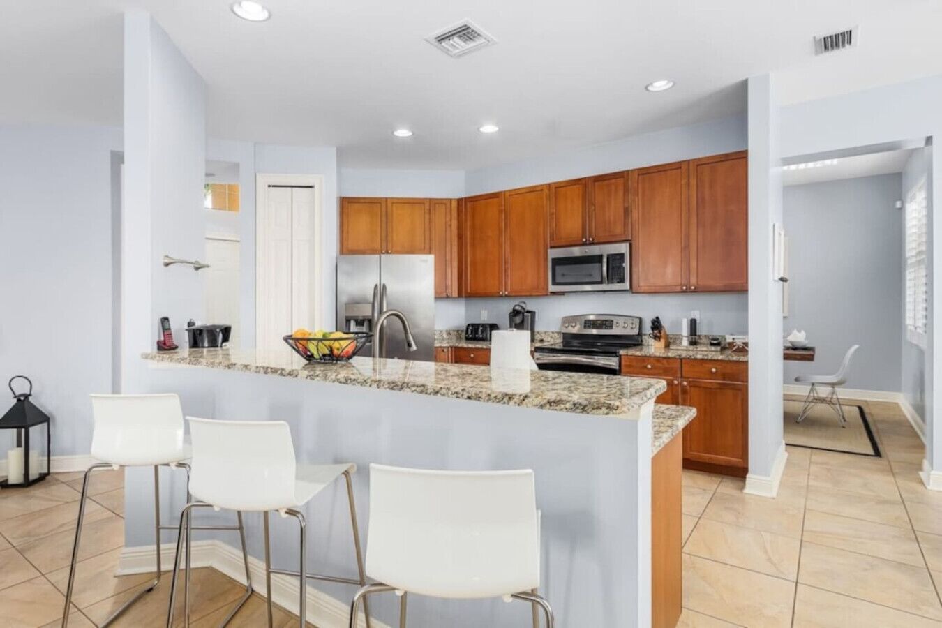 A modern kitchen with granite countertops, wooden cabinets, a stainless steel refrigerator, a microwave, and a gas stove. There are three white bar stools and a small island with a bowl of fruit.