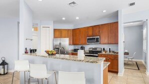 A modern kitchen with granite countertops, wooden cabinets, a stainless steel refrigerator, a microwave, and a gas stove. There are three white bar stools and a small island with a bowl of fruit. - Villa American Beauty (Cape Coral)