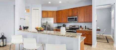 A modern kitchen with granite countertops, wooden cabinets, a stainless steel refrigerator, a microwave, and a gas stove. There are three white bar stools and a small island with a bowl of fruit.