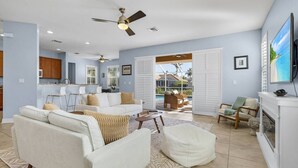 A spacious living room with a white sofa set, a coffee table, and a fireplace. There is a kitchen area with wooden cabinets and a dining table. The room has a large window and a door leading to a balcony with outdoor furniture. - Villa American Beauty (Cape Coral)