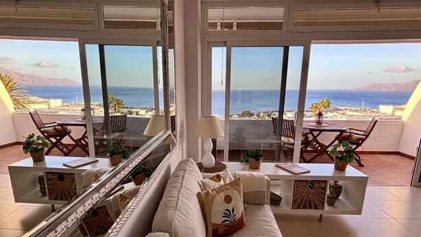 Ein geräumiges Wohnzimmer mit großem Fenster und Blick auf die Küste, einer Couch, einem Couchtisch und einem Essbereich mit Stühlen.
