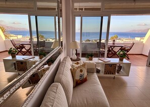 A spacious living room with a large window overlooking a coastal view, a sofa, a coffee table, and a dining area with chairs.