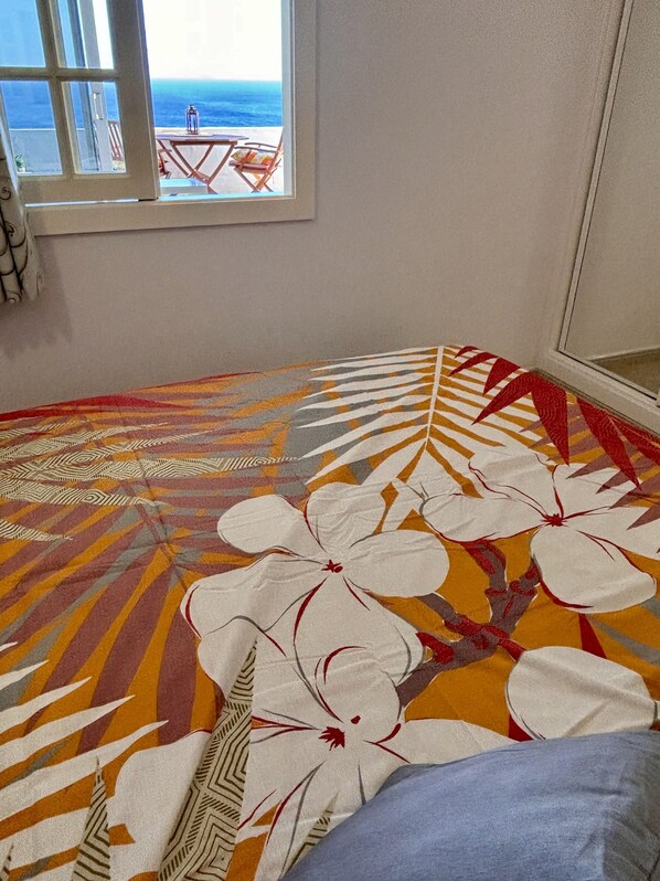 A room with a large window overlooking the sea, a patterned rug, and a bed with a blue pillow.