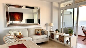 Ein modernes Wohnzimmer mit einer Couch, einem Esstisch mit roter Tischmatte, einem großen Spiegel, einer Lampe und Meerblick durch eine Glastür.