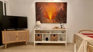 Ein Wohnzimmer mit Fernseher, einer weißen Kommode mit Korbwäsche und einem Gemälde, sowie einem Tisch mit rotem Tischläufer.