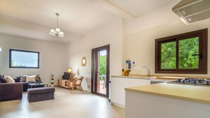 Ein modernes Wohnzimmer mit einer Couch, einem Hocker und einem Essbereich mit Tisch und Stühlen. Eine Küche mit Fenster und Spüle ist ebenfalls zu sehen.