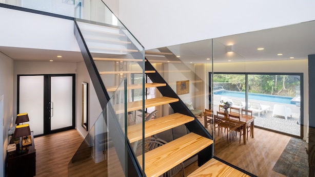 Un interior moderno con una escalera de vidrio, suelo de madera, zona de comedor con asientos exteriores y vista a una piscina.