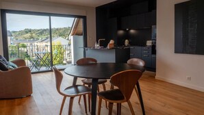 A modern kitchen with dark cabinetry and a large island, a dining area with a round table and chairs, and a balcony with a view of houses and greenery. - Appartement Spacieux Centre Ville (Espalion)