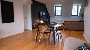 A modern living room with a dining area, a sofa, and a TV mounted on the wall. - Appartement Spacieux Centre Ville (Espalion)
