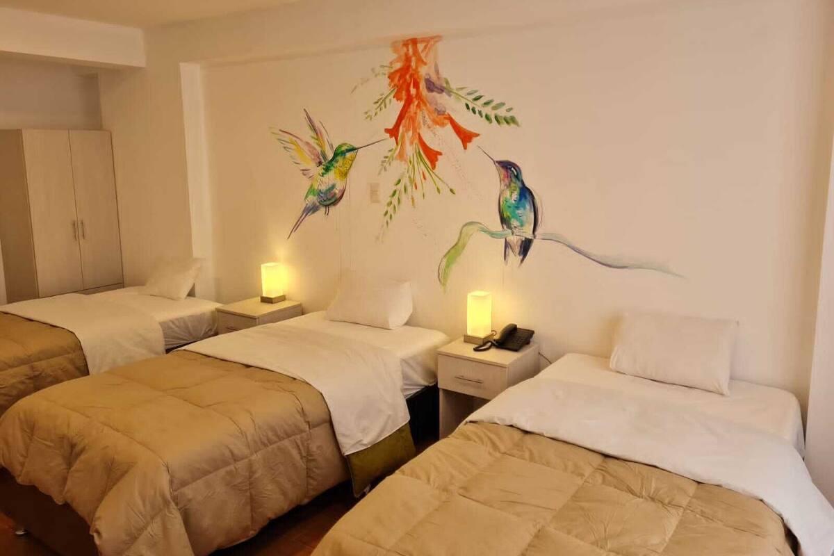 A hotel room with two beds, each with a beige duvet and white pillows. There are bedside tables with lamps and a phone. The walls feature a mural of colorful birds.