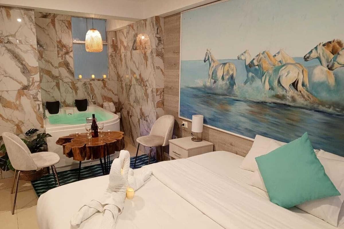 A hotel room with a bed, a painting of horses, a desk with a chair, a small table with a bottle, and a bathtub.