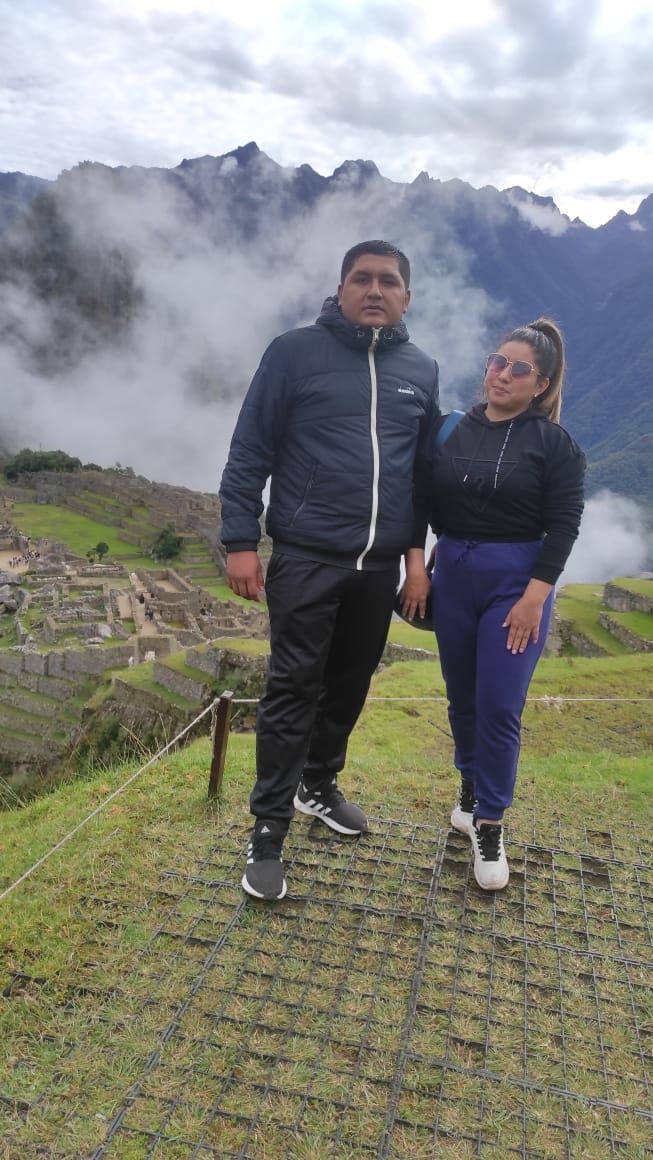 Un hombre y una mujer de pie en una zona cubierta de hierba, con un paisaje montañoso al fondo.
