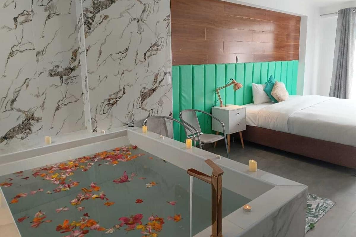 A modern hotel room with a marble-patterned wall, a wooden headboard, a bed with green and white bedding, a small desk, and a bathtub with floating flower petals.