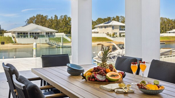A dining table with a fruit platter, glasses, and cutlery, overlooking a water body and buildings.