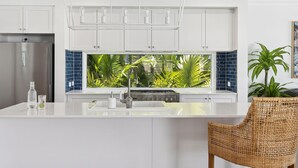 A modern kitchen with white cabinets, a large window with a view of tropical plants, a stainless steel refrigerator, and a wicker chair.