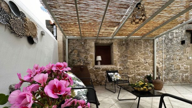 A covered outdoor seating area with stone walls, a table, chairs, and a potted plant with pink flowers. - Afroditi,2 bedrooms,pool,Margarites,Rethymno,Crete (Pigouniana)
