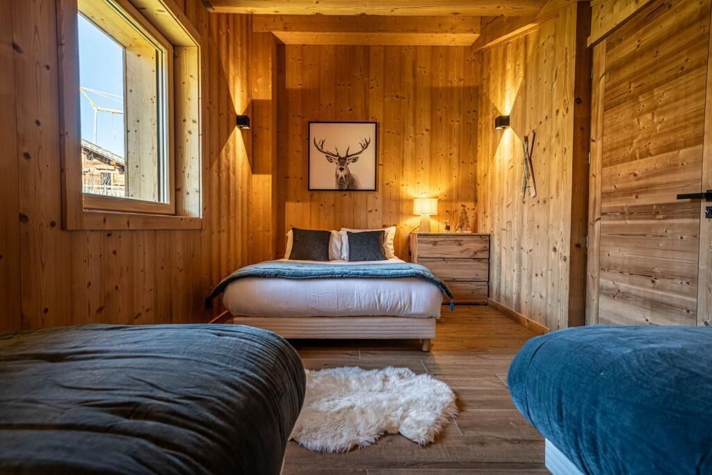 A cozy wooden cabin bedroom with a bed, a small bedside table, a lamp, and a framed deer head picture on the wall.