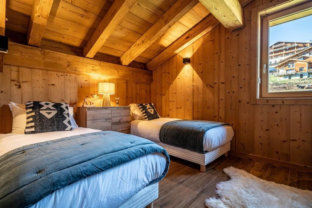 Une chambre confortable, aménagée dans un chalet en bois, avec deux lits, une table de chevet, une lampe et une fenêtre donnant sur un immeuble.
