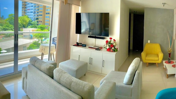 A modern living room with a large flat-screen TV, grey sofas, a yellow armchair, and a view of a cityscape through large windows.