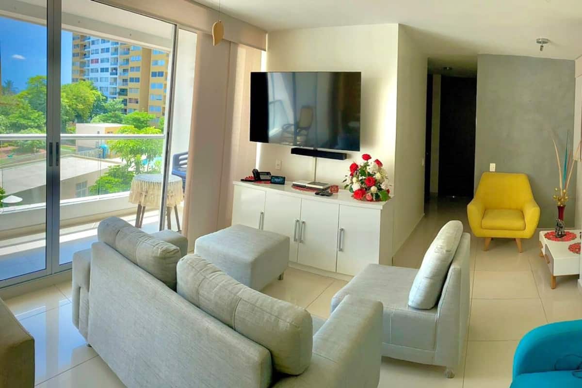 Een moderne woonkamer met een grote flatscreen televisie, grijze banken, een gele fauteuil en een stadsgezicht door grote ramen.