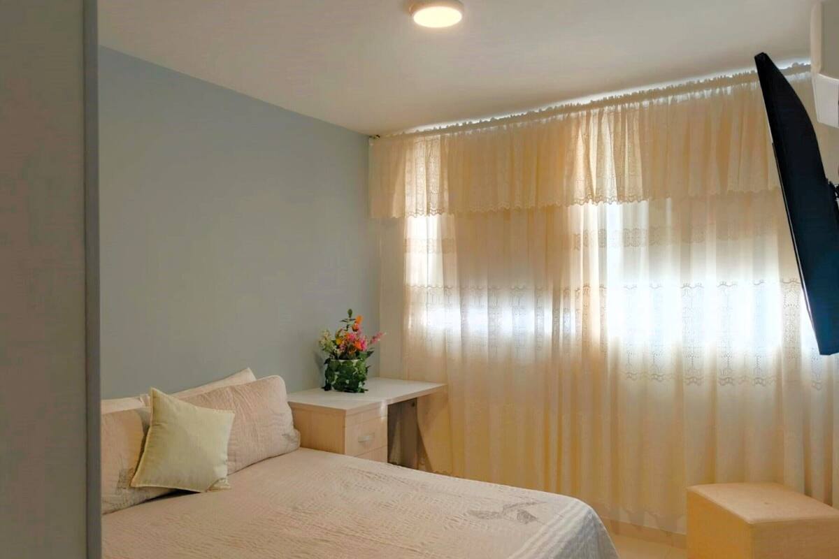 Een slaapkamer met een bed, een klein nachtkastje met een bloevas en een raam met dunne gordijnen.