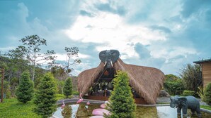 A large elephant statue in a garden with a pond, trees, and a thatched-roof structure. - Selva Ecospa Wellness Stay (Guatapé)