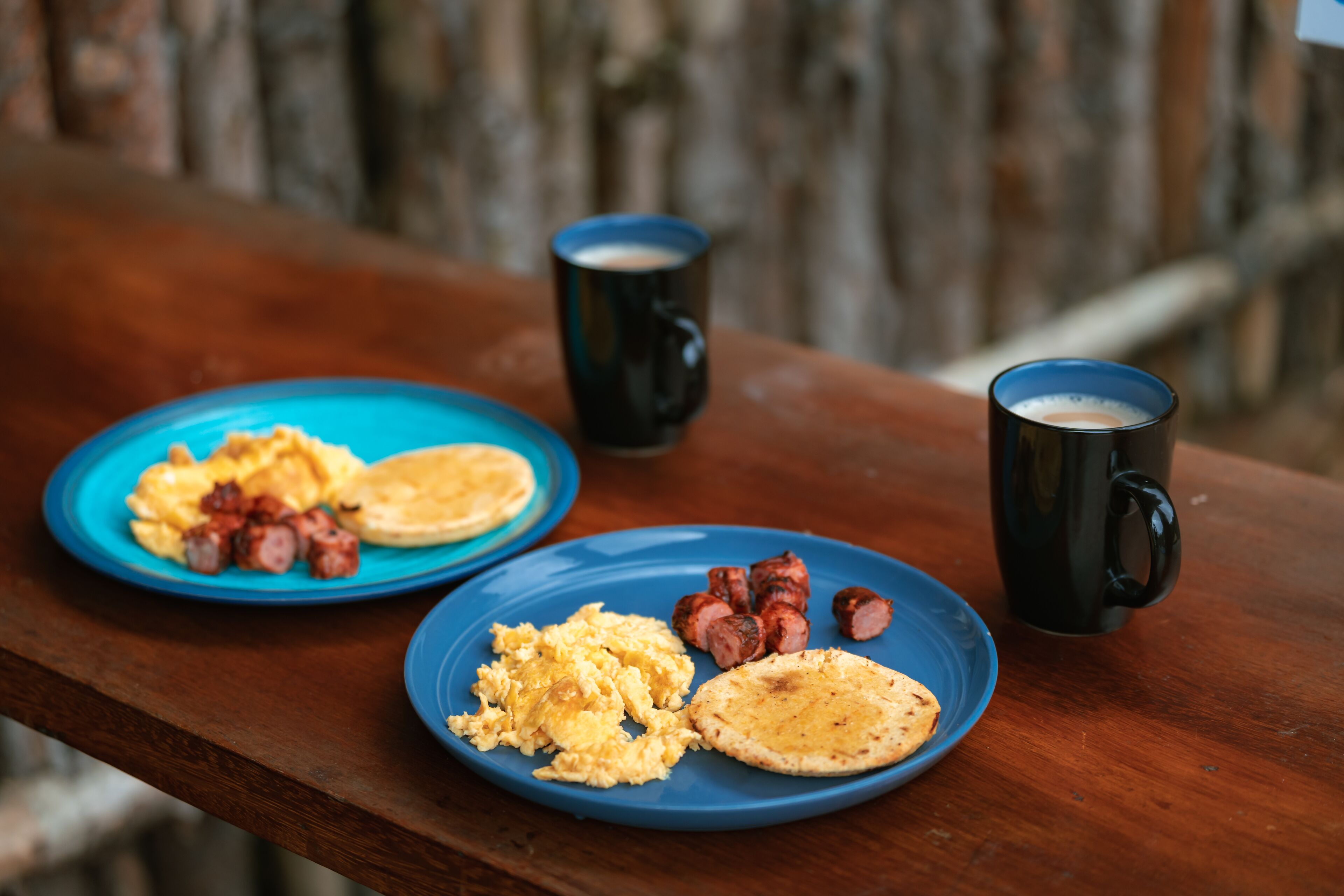 Two blue plates with scrambled eggs, sausage, and a flatbread on a wooden table.