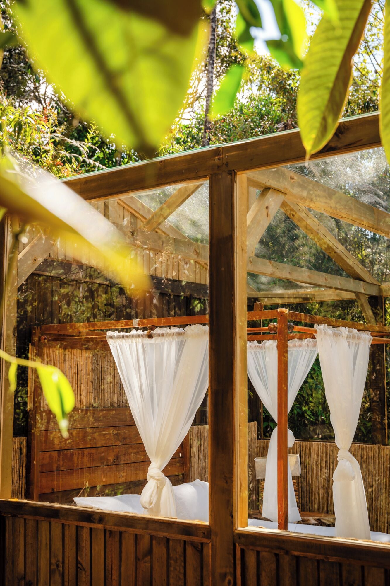 Une structure en bois surmontée d’une verrière, avec des rideaux blancs et un lit à baldaquin.