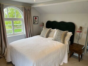 A bedroom with a large bed, a window with curtains, a bedside table, and a wall-mounted lamp.
