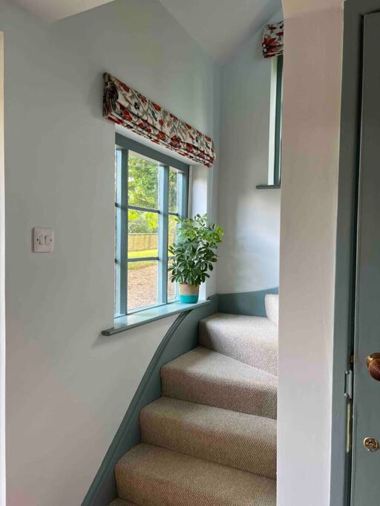 Una escalera con una planta en el rellano, una ventana con un adornos estampados y una puerta a la derecha.