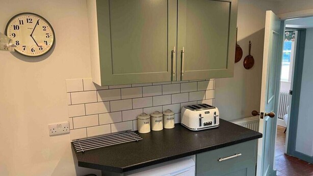 A compact kitchen with green cabinets, a black countertop, a toaster, and a wall clock. - Luxury Cottage with Garden (Tewkesbury)