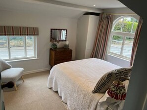 A bedroom with a large bed, a chair, a dresser, a mirror, and a window with curtains. - Luxury Cottage with Garden (Tewkesbury)