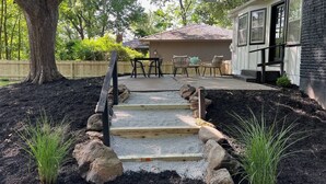 Un chemin mène à un espace patio aménagé avec une table et des chaises, le tout entouré d’une clôture en bois et d’un arbre.