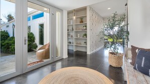 Un salon moderne avec une porte vitrée donnant sur l’extérieur, une bibliothèque intégrée, une grande plante en pot et un fauteuil en osier.