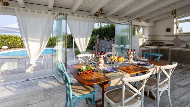 An outdoor dining area with a pool view, a glass partition, and a kitchen in the background. - Villa Alexandra - Villa with pool (Ispica)