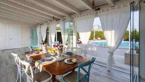 A covered outdoor dining area with a pool view, wooden tables, white chairs, and a glass door leading to the pool. - Villa Alexandra - Villa with pool (Ispica)