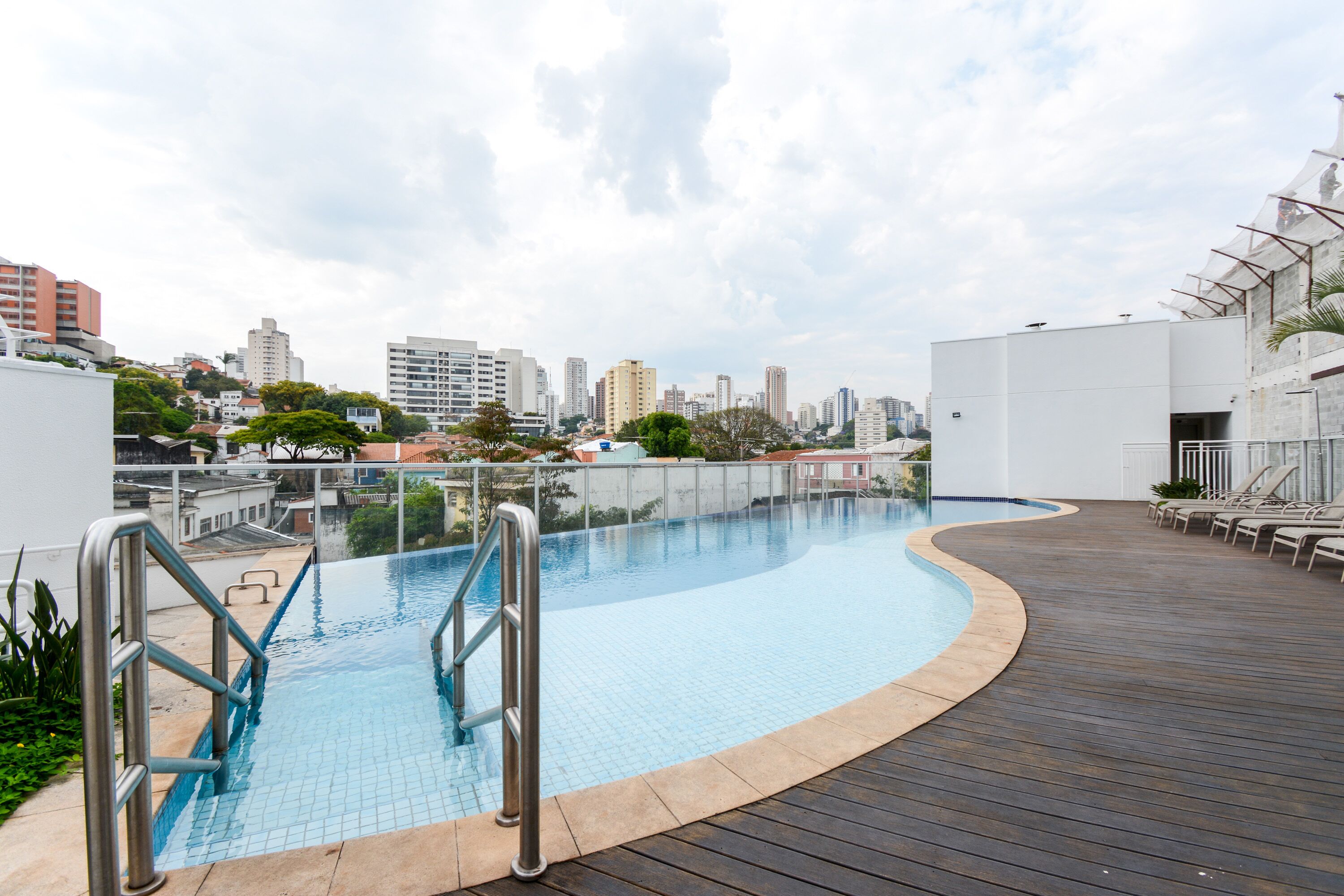 A rooftop swimming pool with a wooden deck and metal railings, surrounded by buildings and a cityscape.