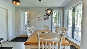 Une salle à manger avec une table en bois et des chaises blanches, de grandes fenêtres et un luminaire suspendu.