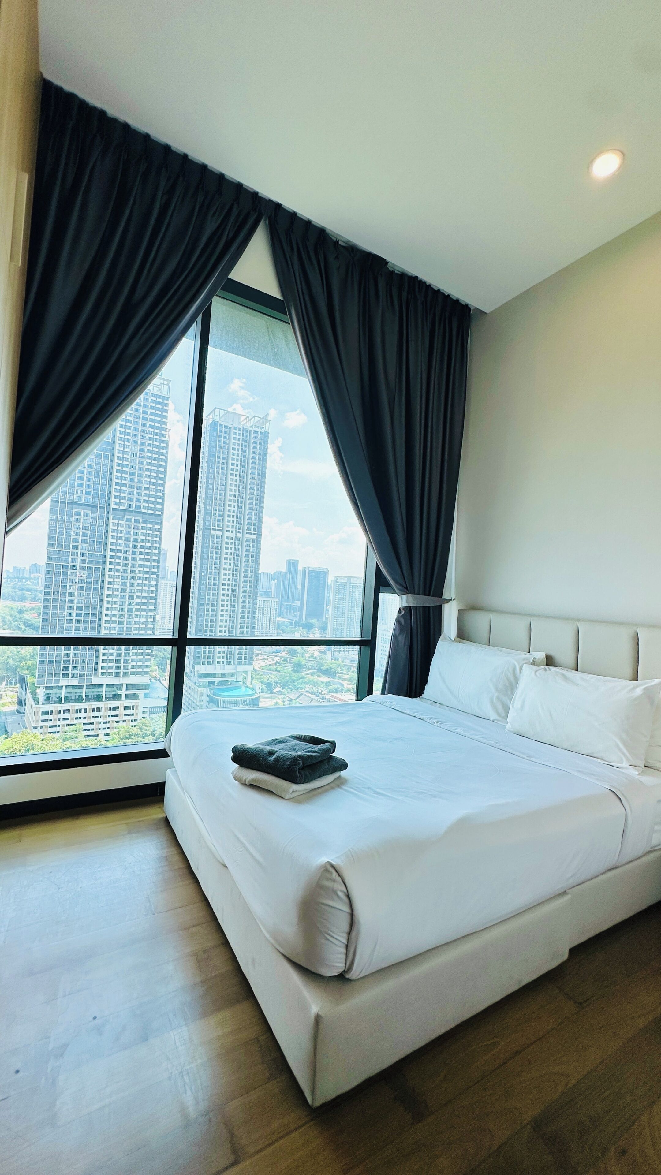 A modern hotel room with a large bed, a view of a cityscape through floor-to-ceiling windows, and a ceiling light.