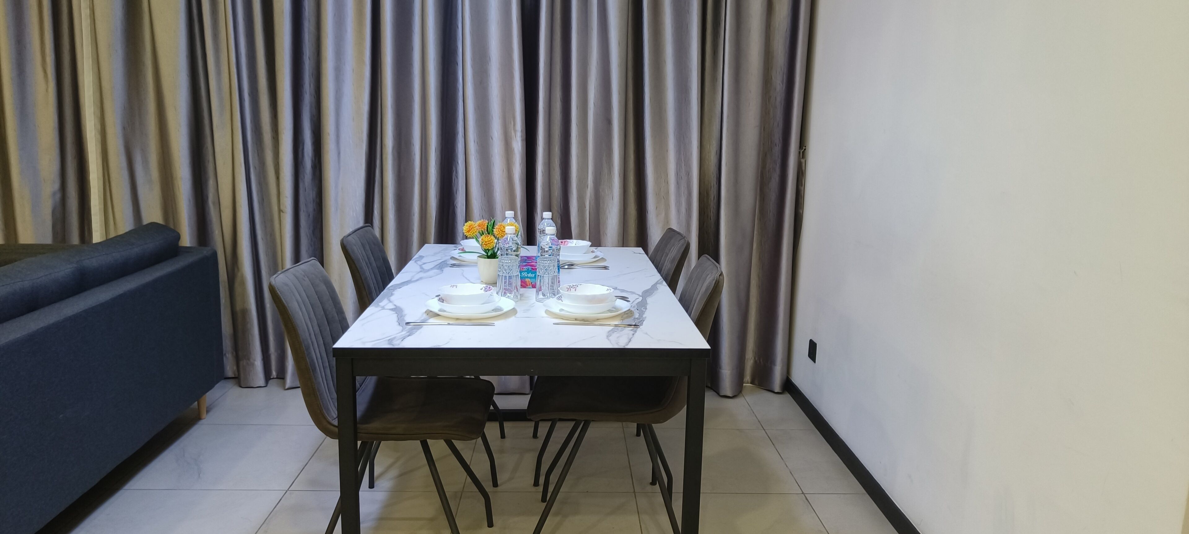 A dining area with a marble table set for four, surrounded by chairs, with a sofa and curtains in the background.