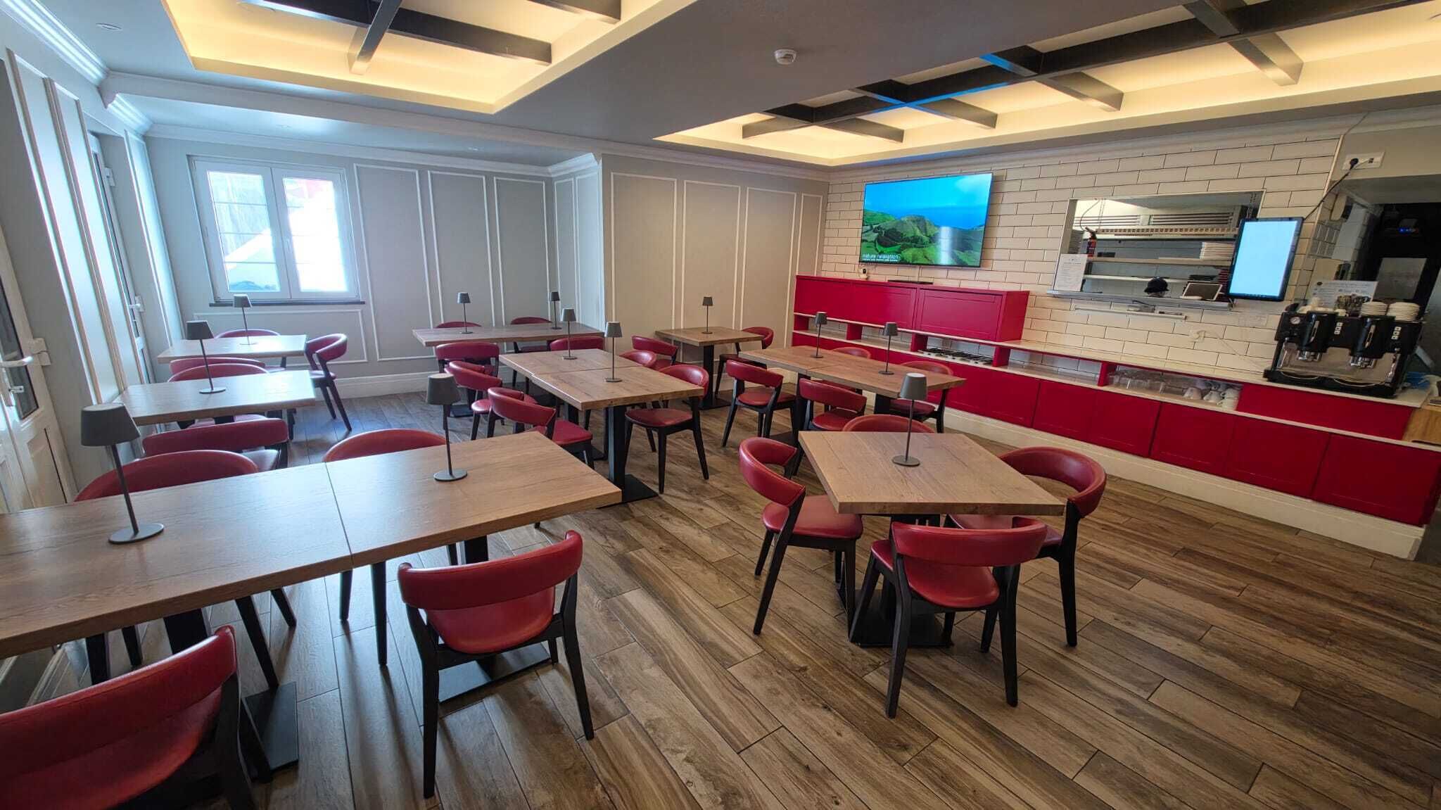 A modern restaurant with wooden tables and red chairs, a kitchen area, and a TV on the wall.