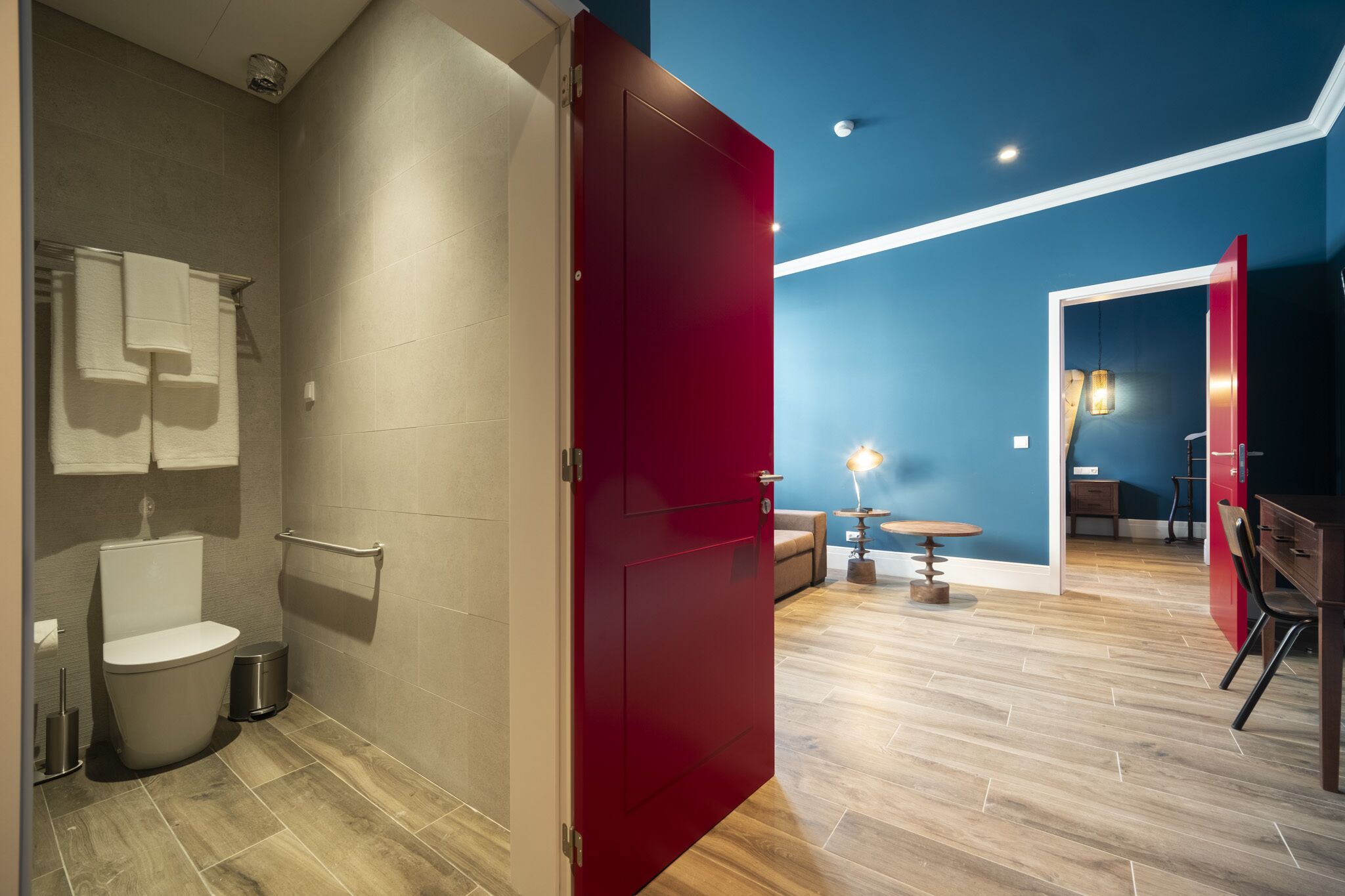 Een moderne badkamer met toilet, handdoekrek en een rode deur naar een woonruimte met houten vloer, een tafeltje en een stoel.
