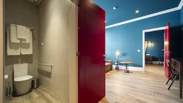 Een moderne badkamer met toilet, handdoekrek en een rode deur naar een woonruimte met houten vloer, een tafeltje en een stoel.