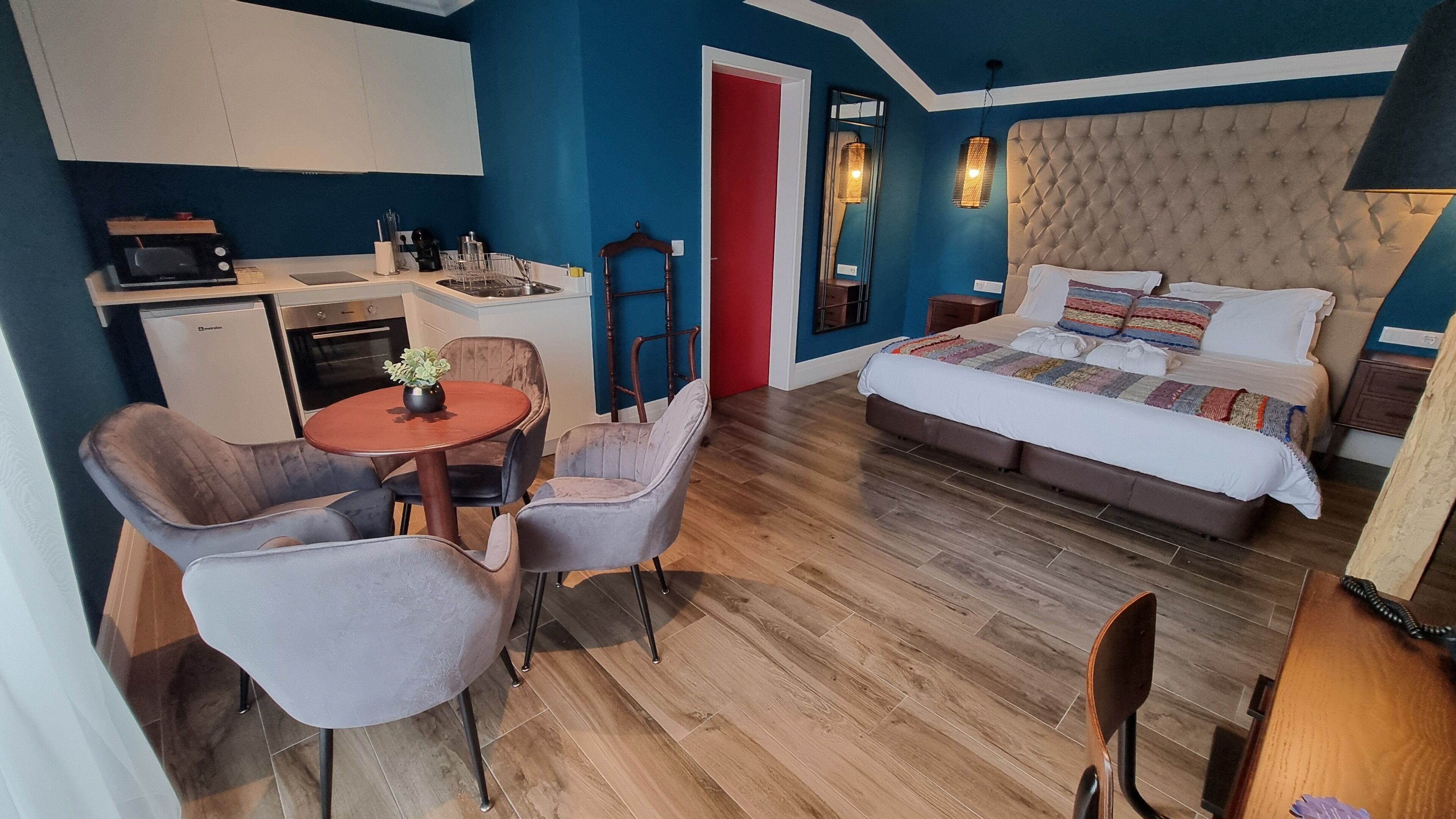 Une pièce compacte comprenant un coin cuisine, un espace repas avec une table et des chaises, et un lit avec une couverture colorée.