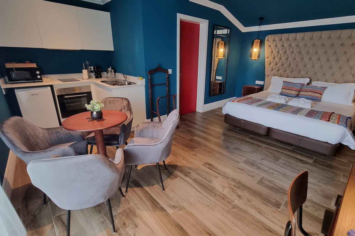 Een compacte kamer met een kitchenette, een eethoek met een tafel en stoelen, en een bed met een kleurrijke deken.