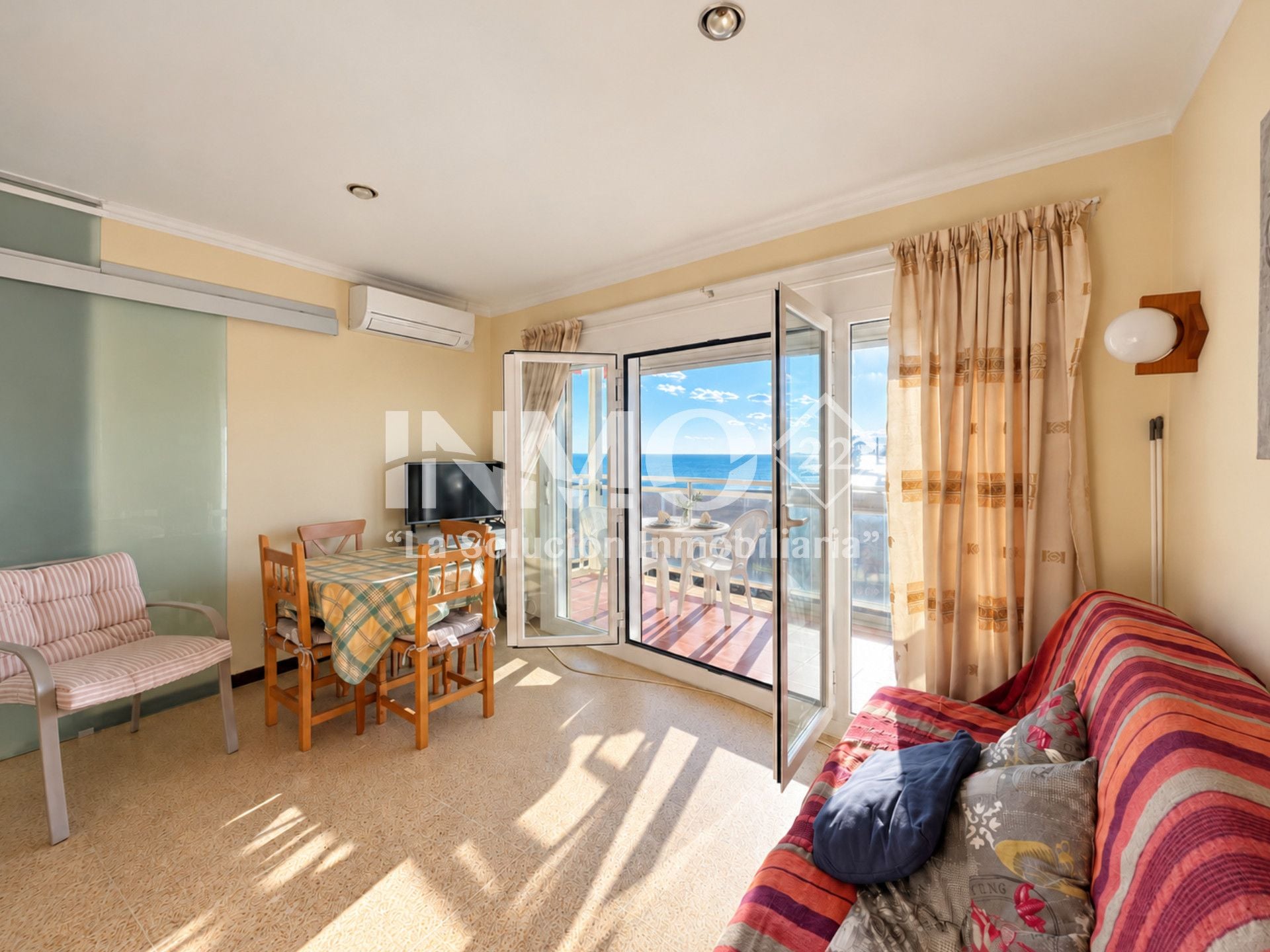 A living room with a sofa, a dining table with chairs, a TV, and a sliding glass door leading to a balcony with a view of the sea.